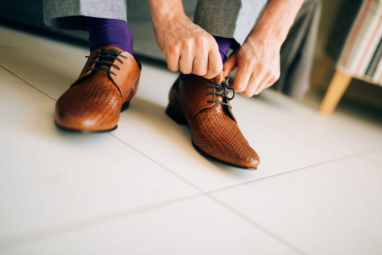 Hands tying the laces of brown woven leather shoes with purple socks on a tile floor