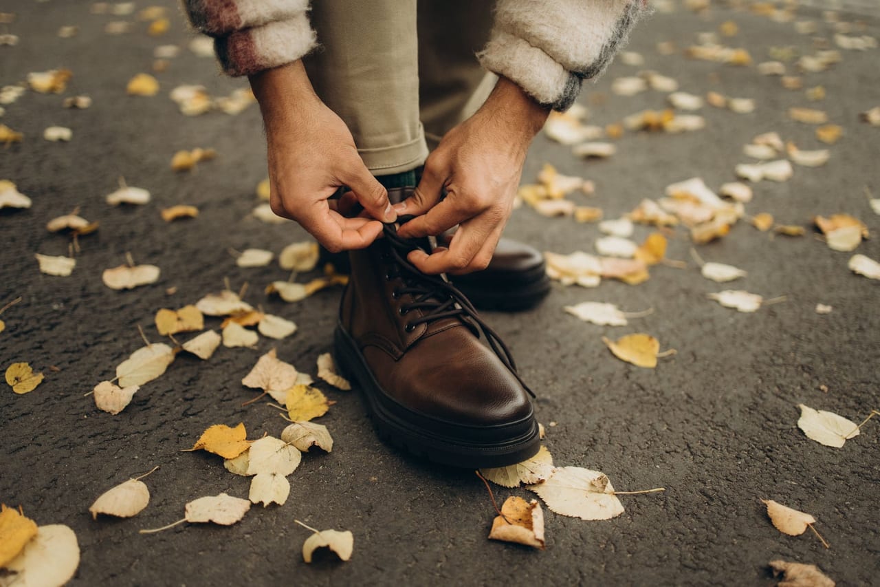 Hands tying the laces of brown leather boots on asphalt scattered with yellow leaves