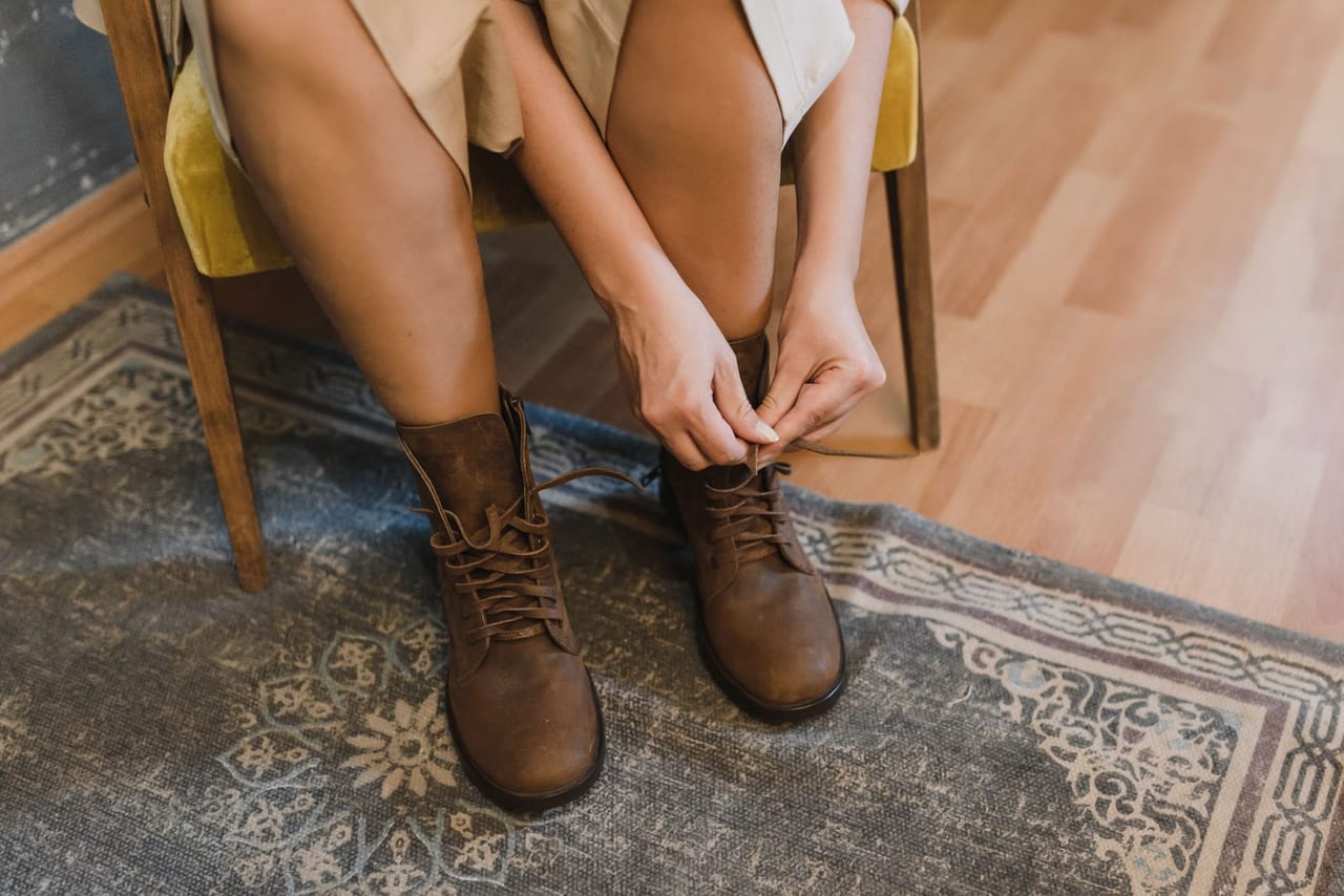 Woman on a yellow chair tying brown leather ankle boots over a patterned rug