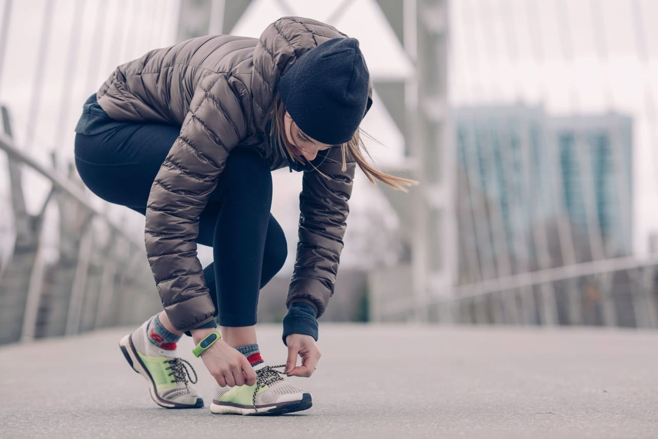 Woman in a puffer jacket crouching to tie her running shoes on a city bridge
