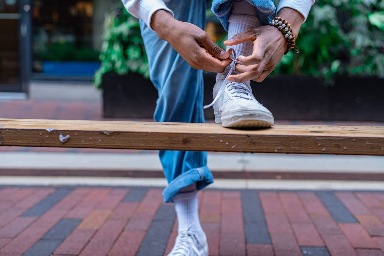 Hands tying the laces of a white sneaker propped on a wooden bench outdoors