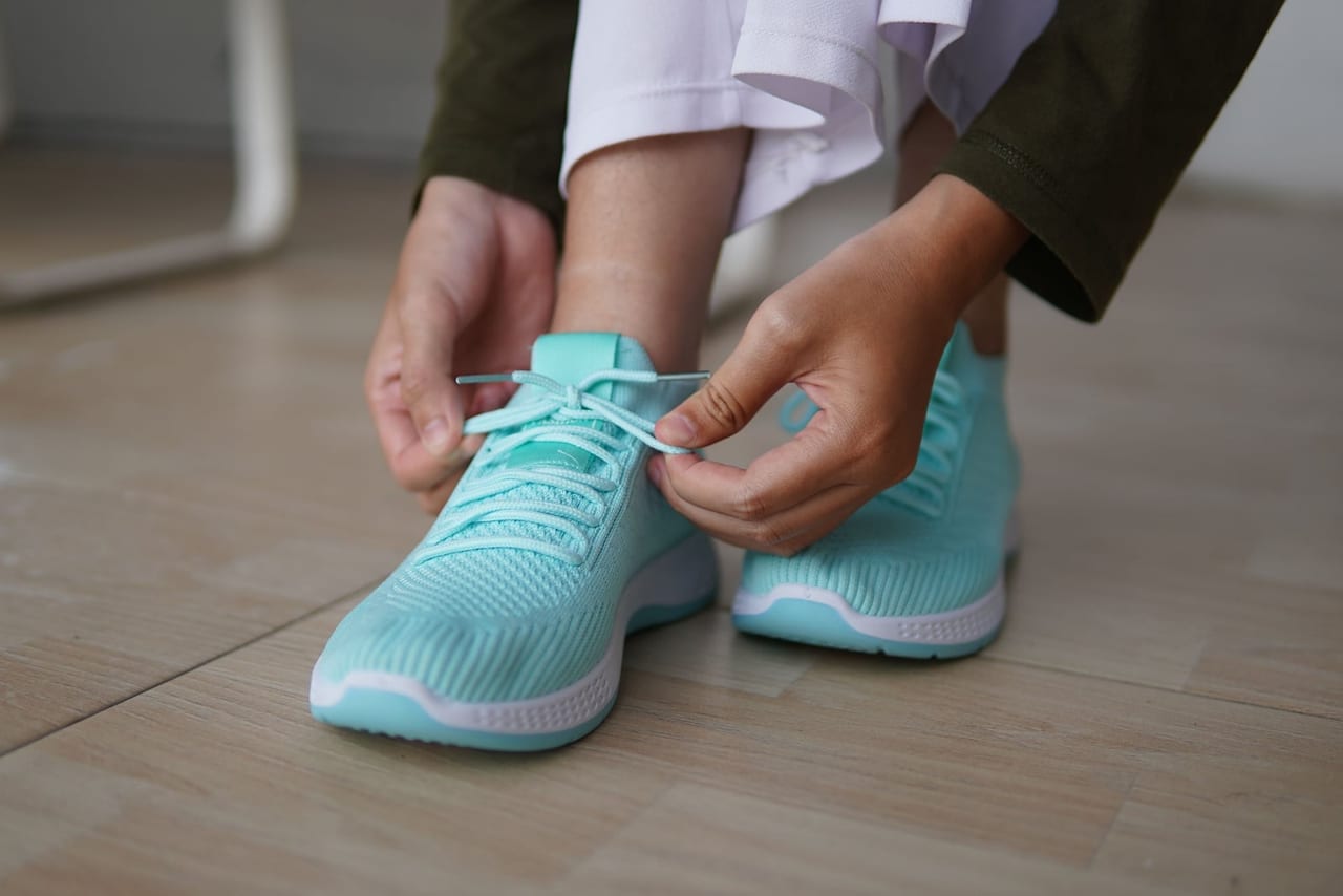 Hands tying the laces of turquoise sneakers on a light-colored floor indoors