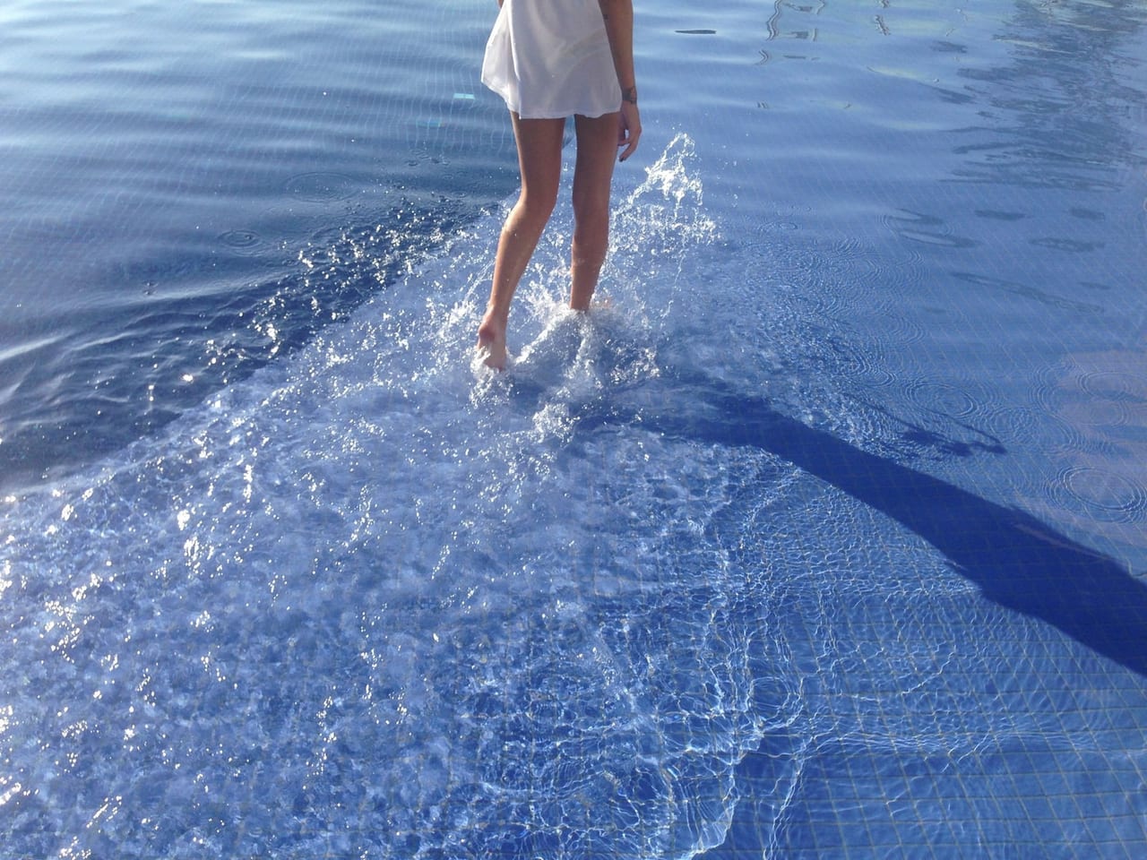 Woman in a short white dress wading through shallow pool water, splashing with each step