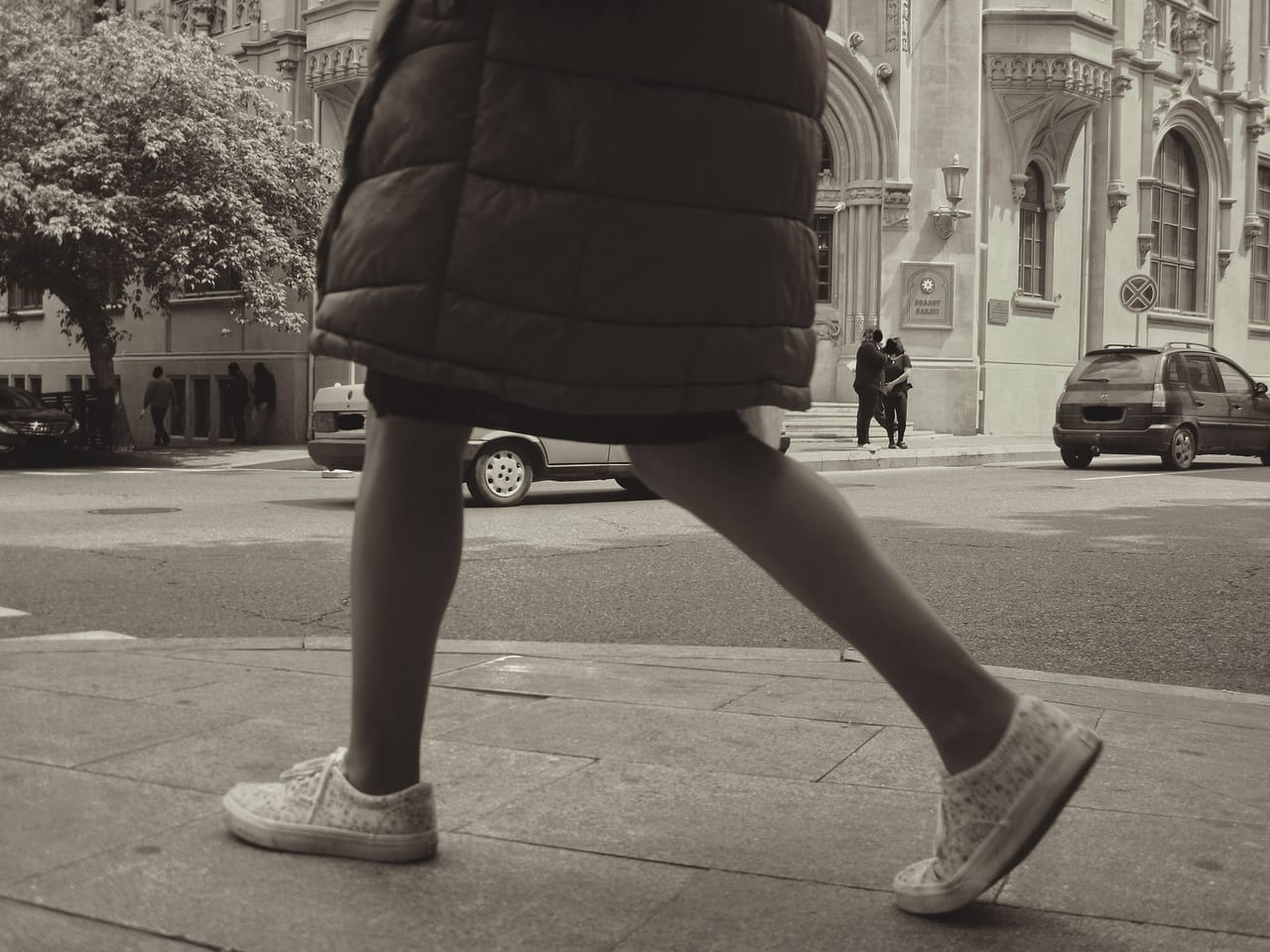 Woman in a quilted coat and canvas sneakers walking along a city sidewalk