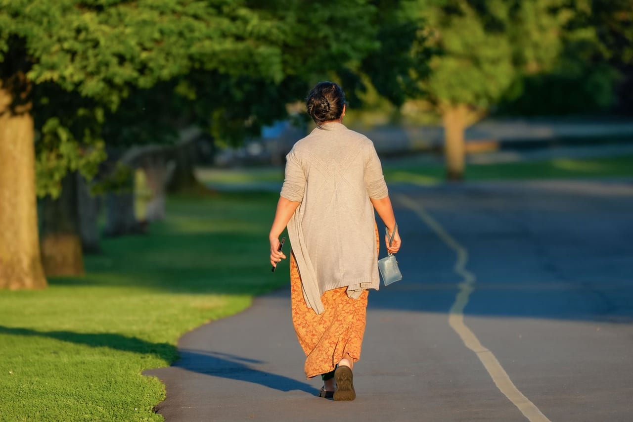 Woman seen from behind walking along a sunlit, tree-lined park path, head up