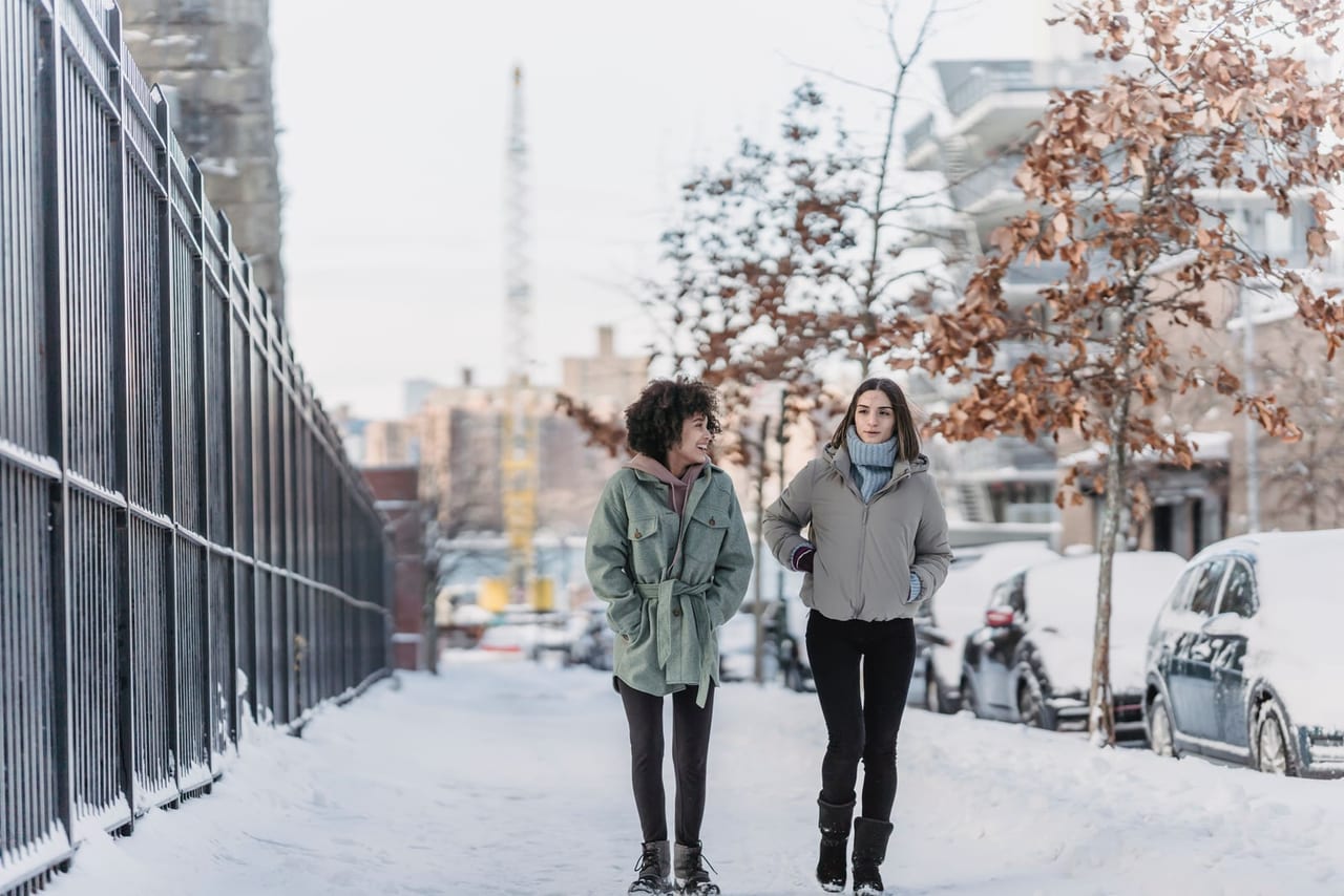 Two women in winter coats and boots walking along a snow-covered city sidewalk