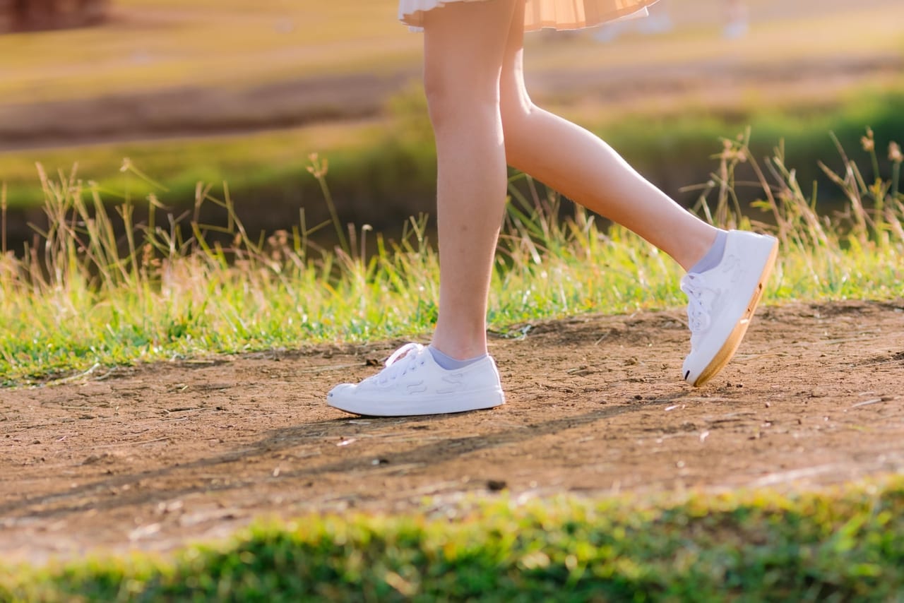 Legs in white sneakers mid-stride on a sunlit dirt path, the back foot pushing off