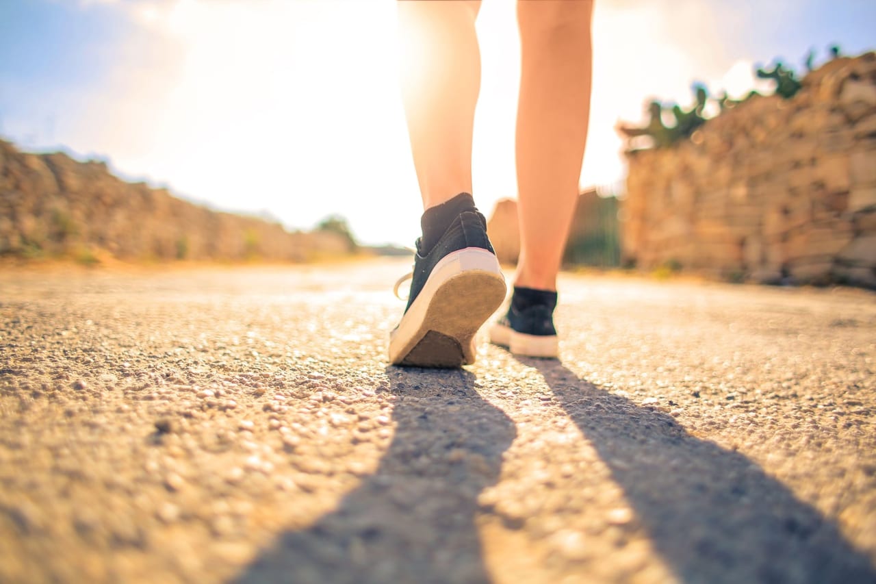 Low view of legs walking along a sunlit country road in black canvas sneakers