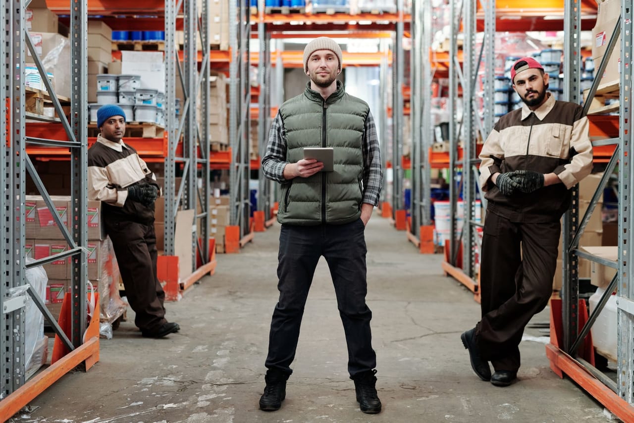 Three warehouse workers standing in an aisle between tall shelves on a concrete floor