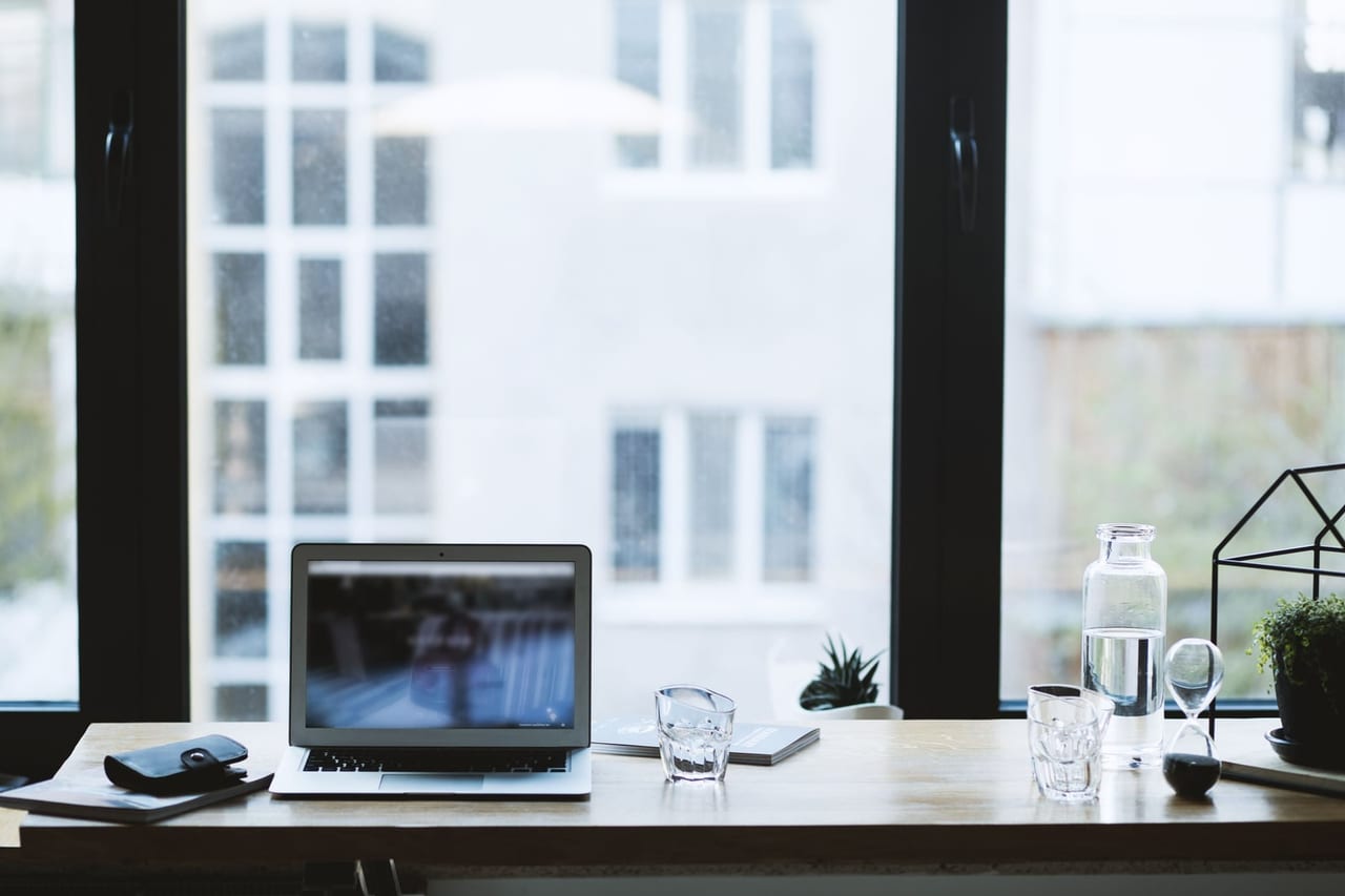 Bottle of water and two glasses on a desk beside a laptop and a bright window
