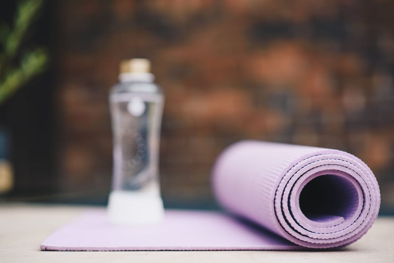 Clear water bottle beside a rolled lilac yoga mat on a pale floor