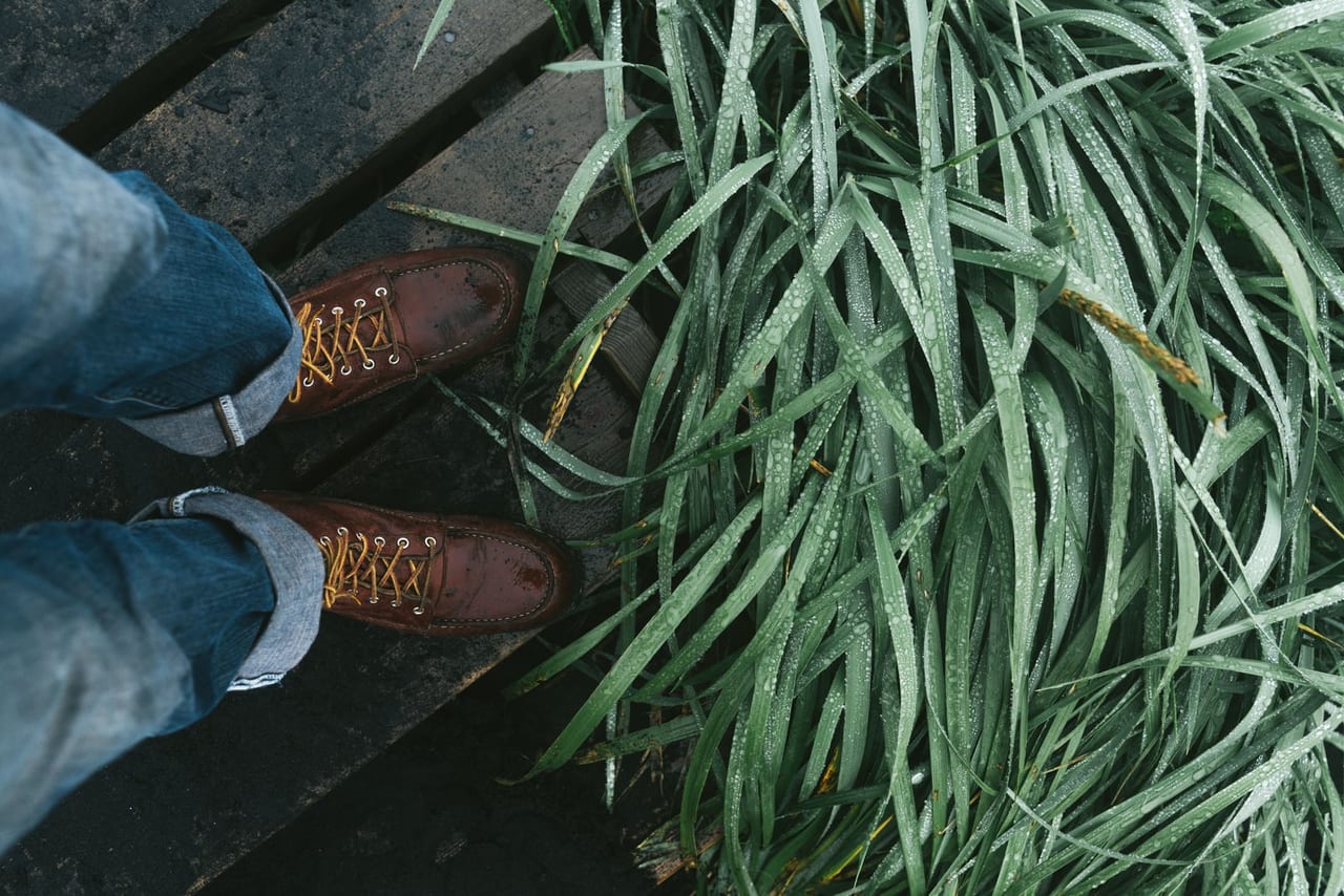 Brown leather lace-up boots, wet with raindrops, on a wooden deck beside dewy grass