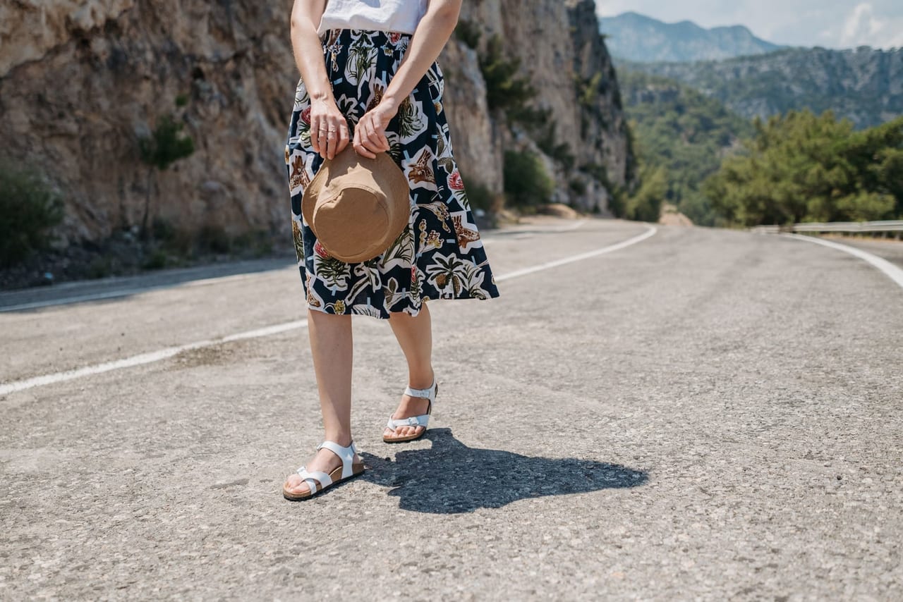 Woman in white open sandals and a floral skirt standing on a quiet sunlit road