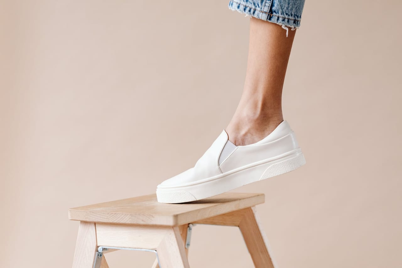 White slip-on shoe in side view, worn with cuffed jeans, resting on a wooden stool