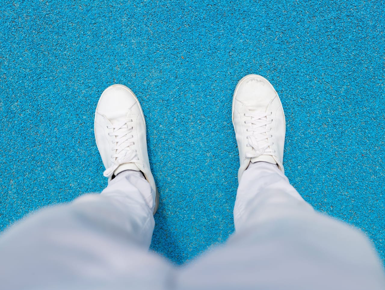 Looking down at scuffed white sneakers and light pants on a textured blue surface