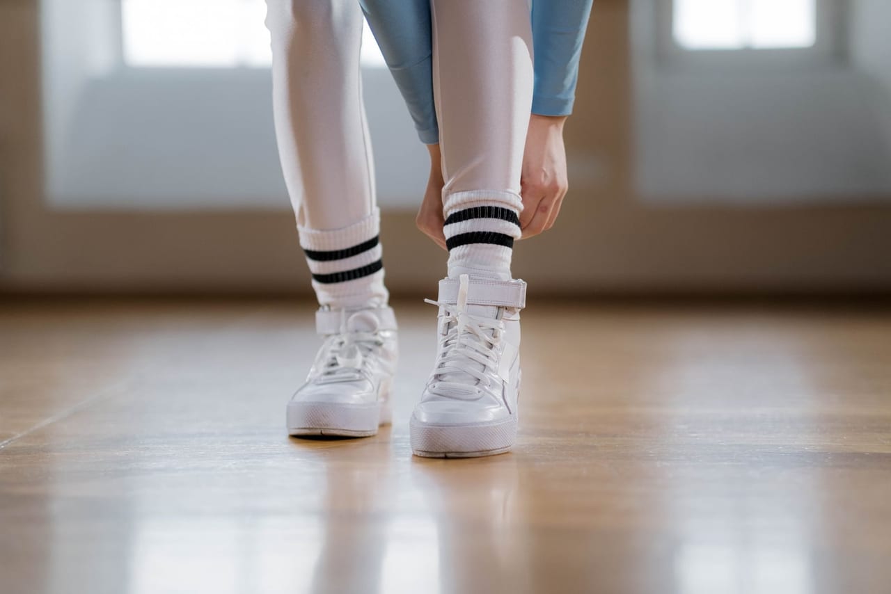 Person in white high-top sneakers and striped socks adjusting an ankle strap on a wooden floor