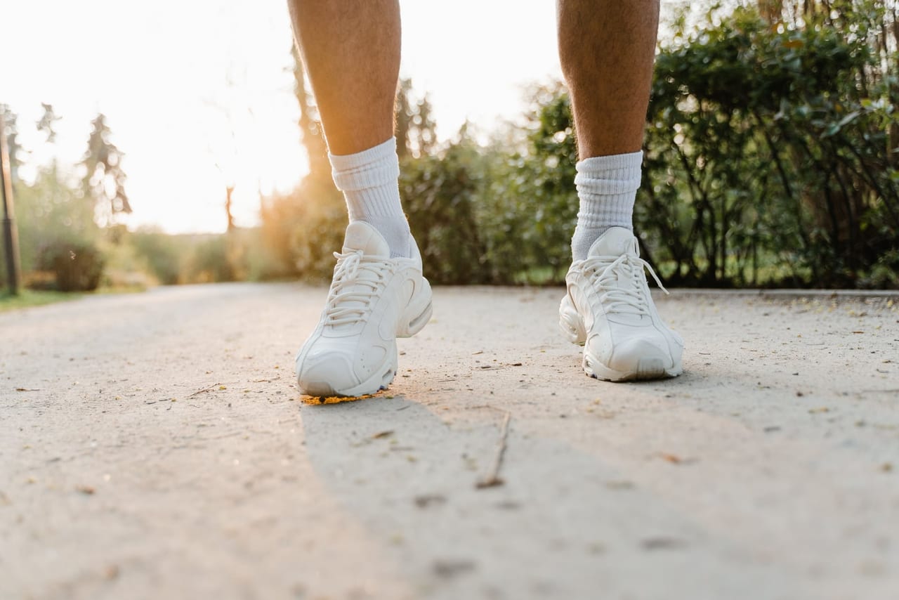 Legs in white sneakers and socks mid-step on a pale gravel path in low sunlight