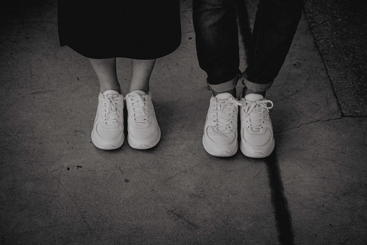Two people in white sneakers standing side by side on rough pavement, in black and white