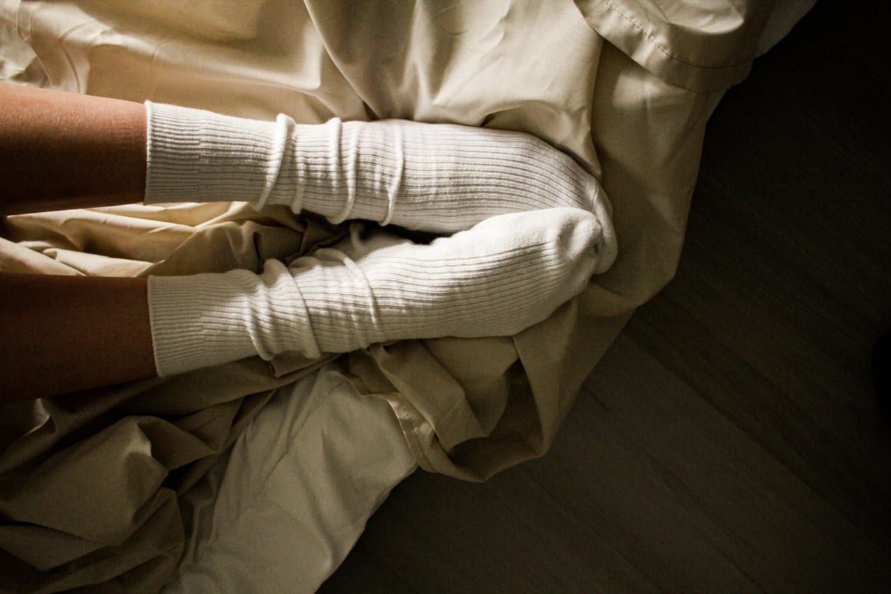 Feet in thick white ribbed socks resting on rumpled beige bedding