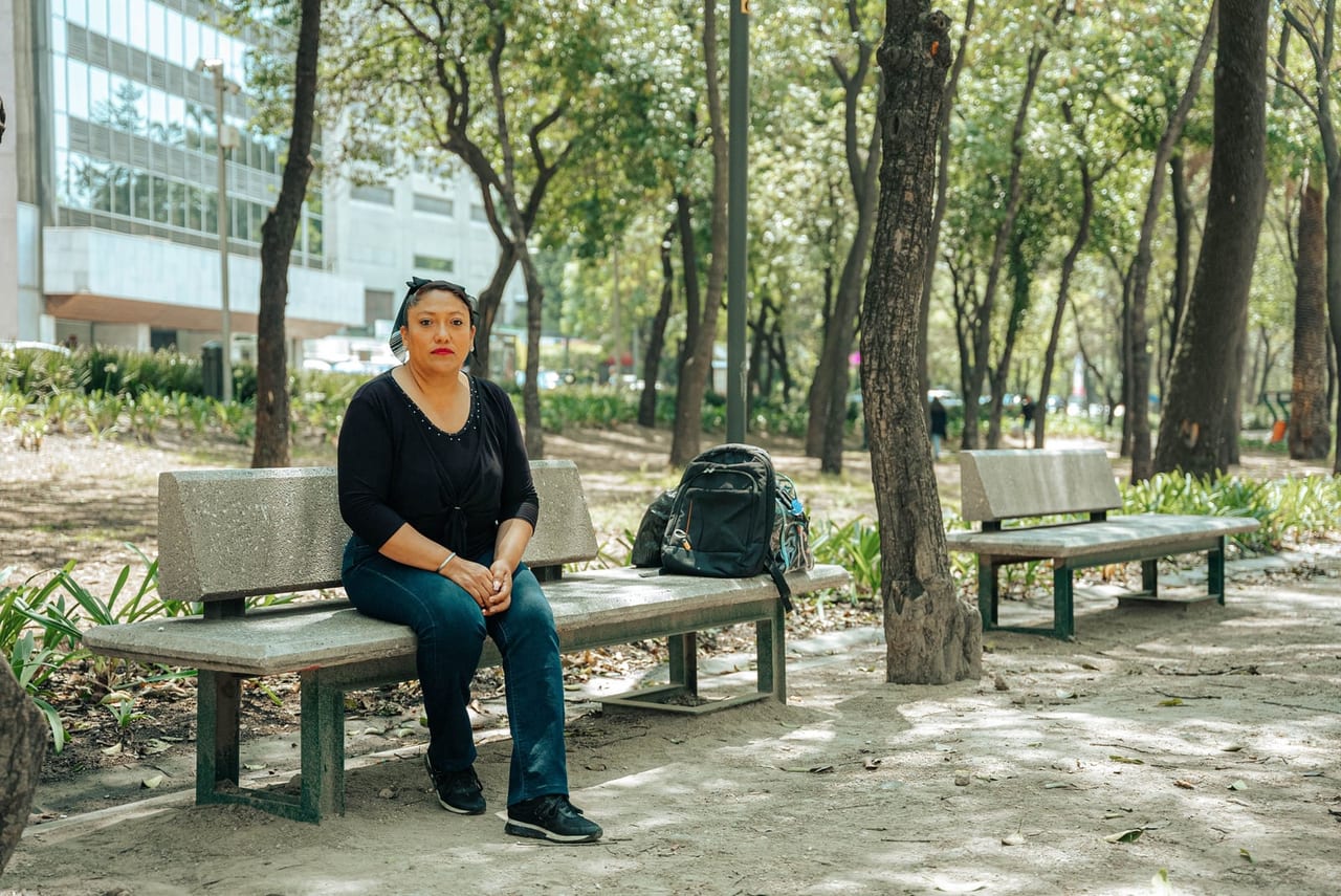 Woman sitting on a stone park bench among trees with a backpack beside her
