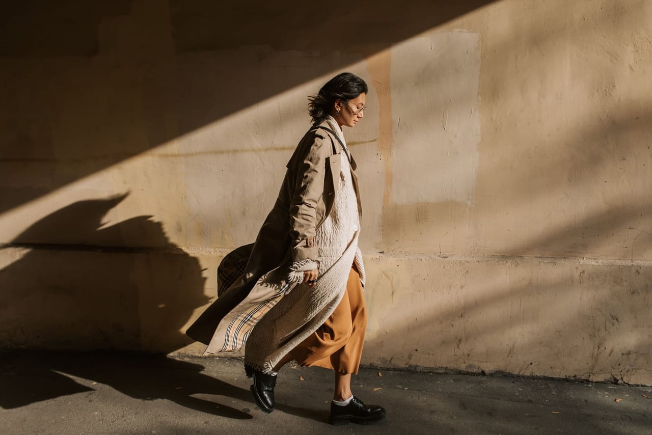 Woman in a trench coat and black lace-up shoes walking past a sunlit wall