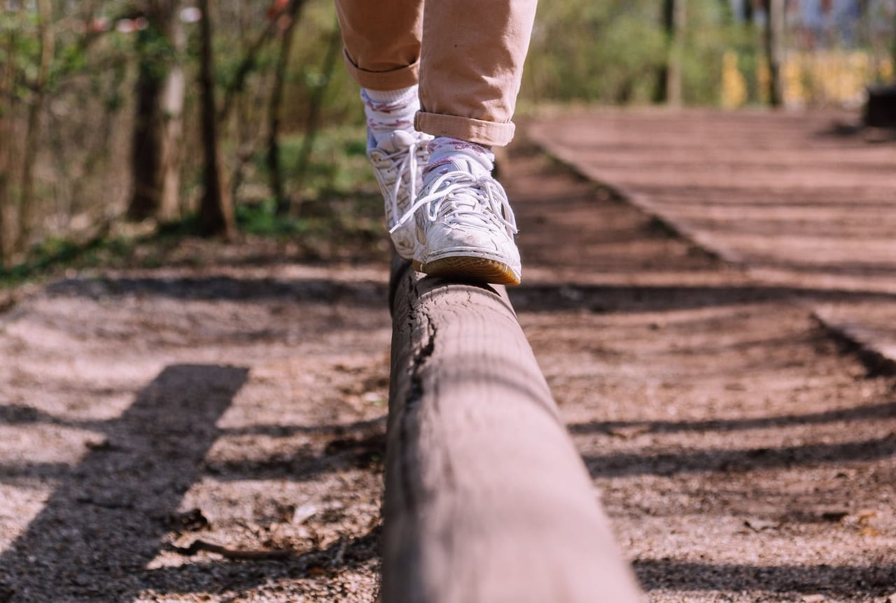 Feet in white sneakers stepping along a narrow wooden beam beside a park path