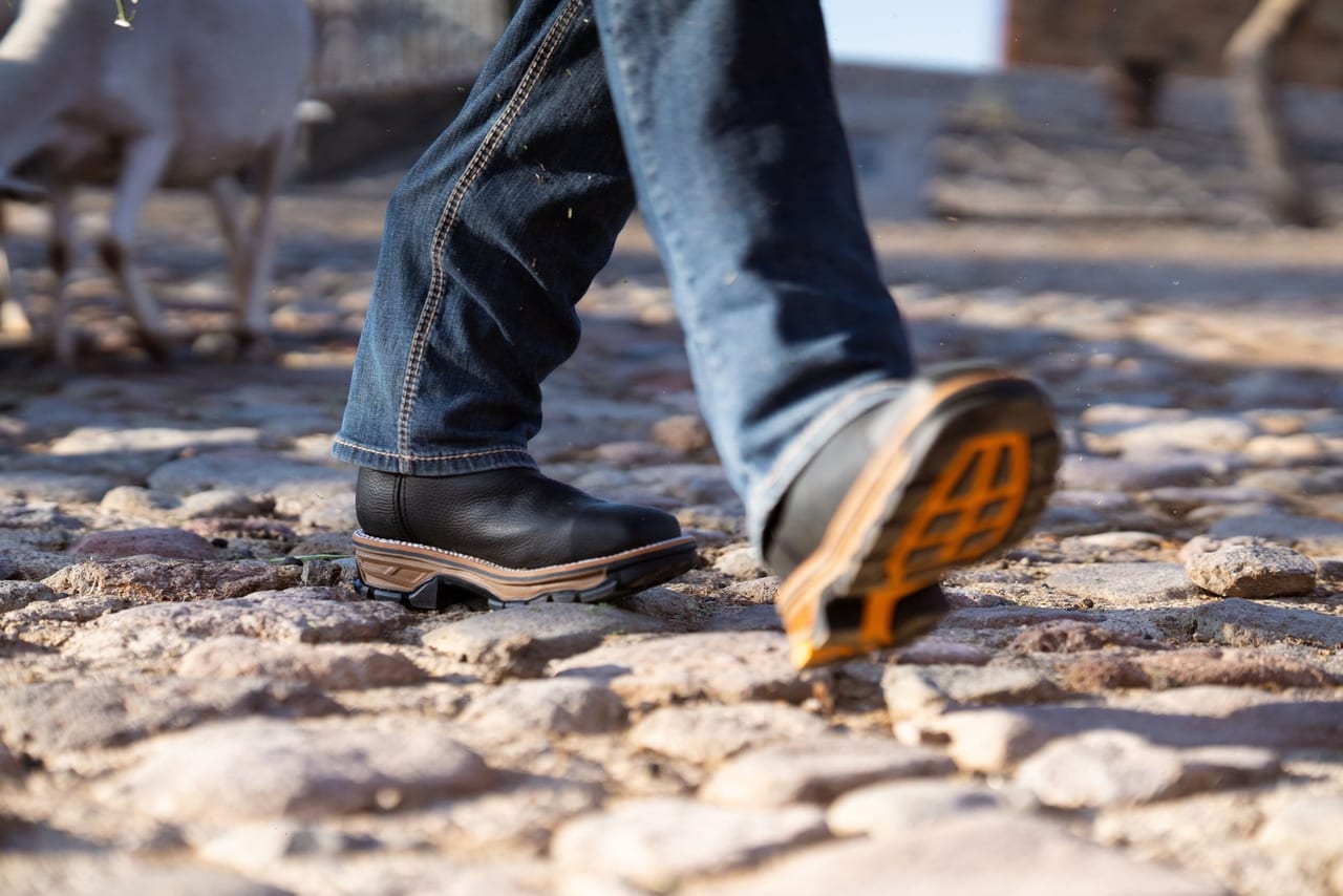 Person in jeans and black work boots walking across sunlit cobblestones, one sole raised