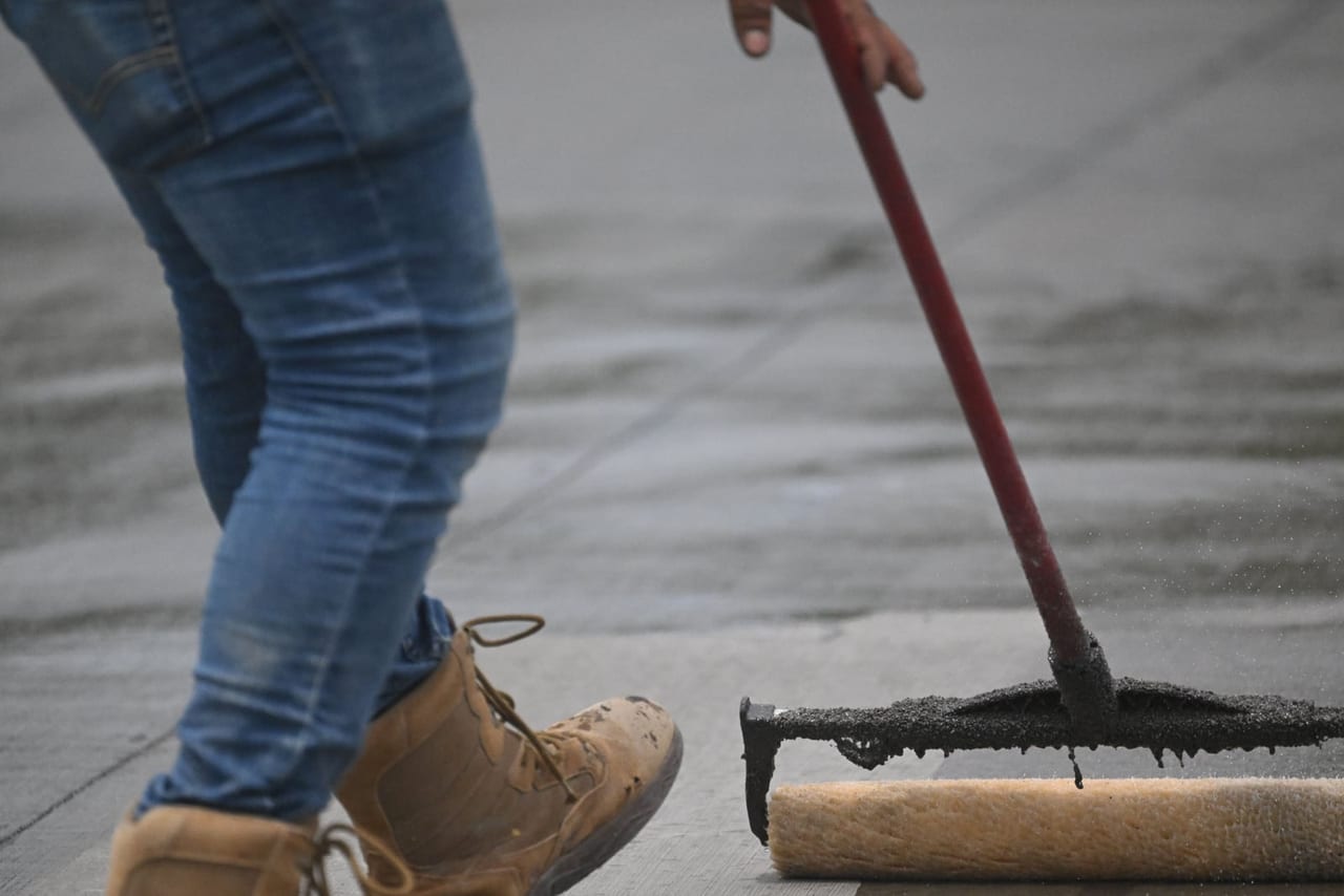 Worker in jeans and tan work boots rolling sealant across a concrete surface