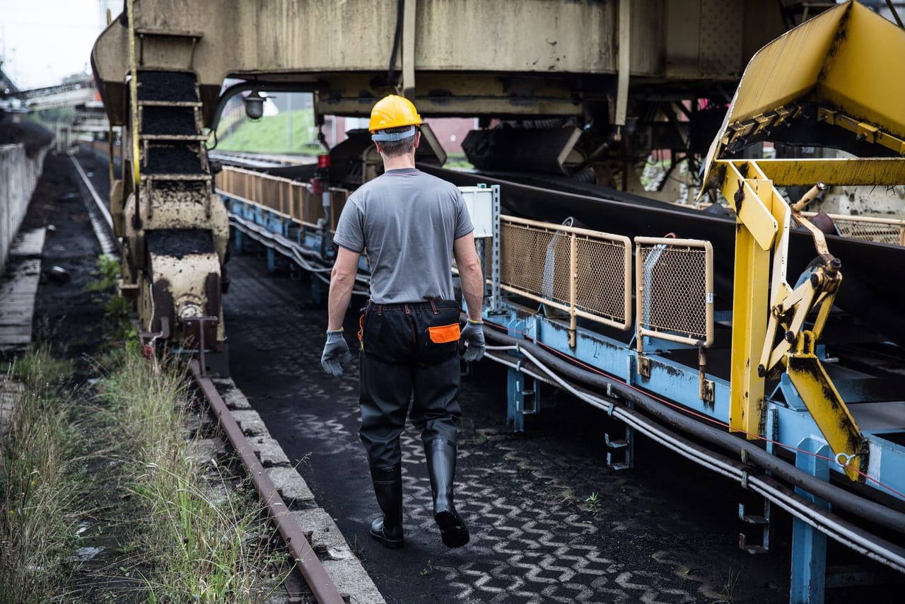 Worker in a hard hat and rubber boots walking beside a conveyor at an industrial site