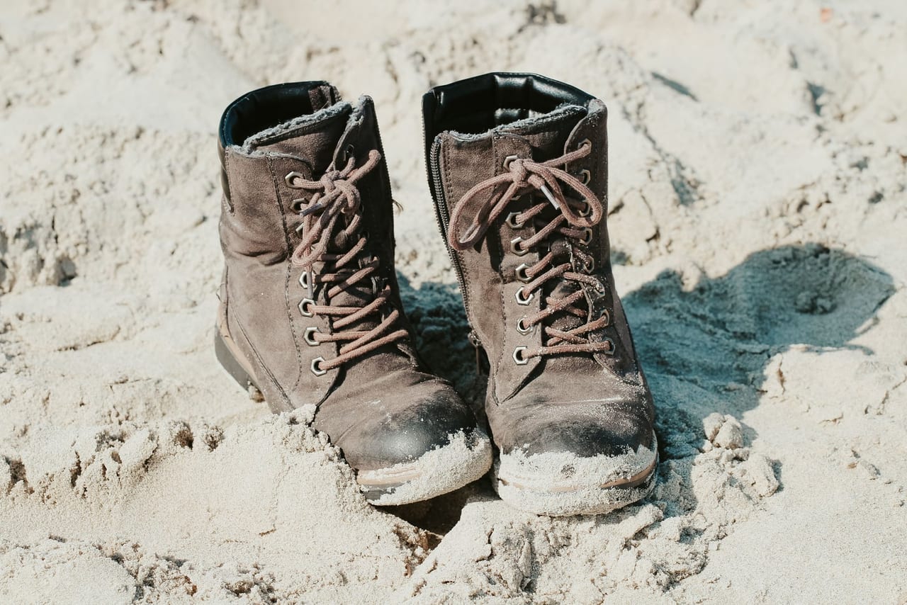Pair of worn brown lace-up boots standing on pale sand with scuffed, sandy toe caps
