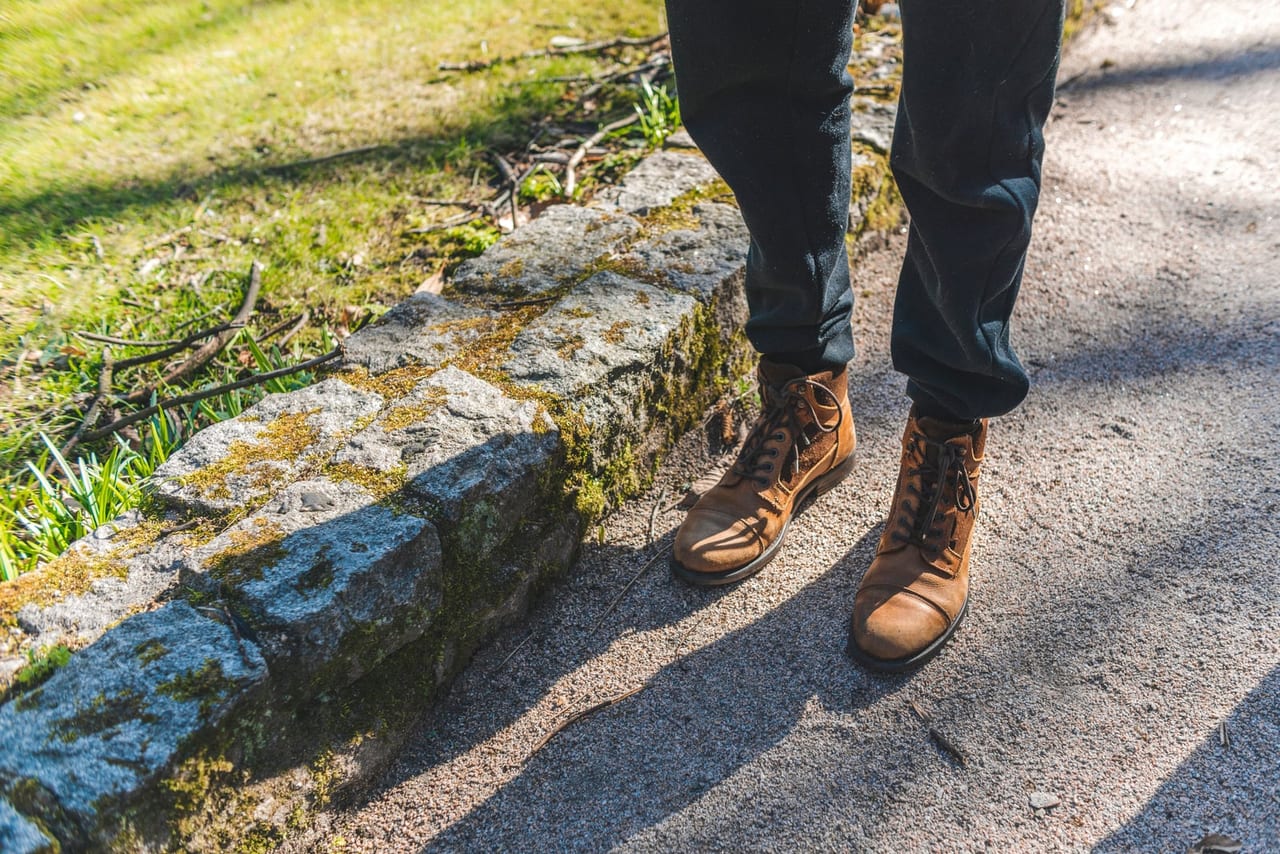 Worn brown leather lace-up boots standing on a sunlit gravel path beside a mossy curb