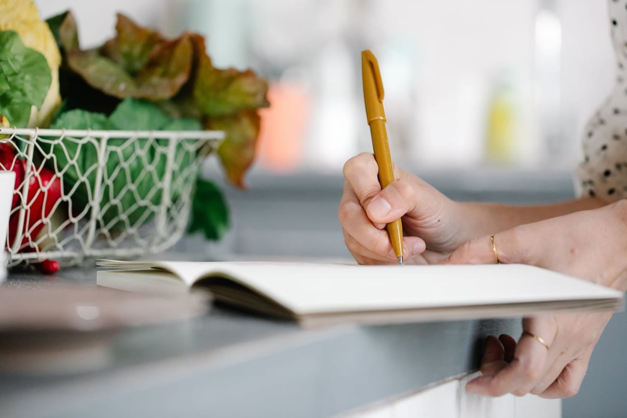 A hand writing in an open notebook on a kitchen counter beside a basket of vegetables