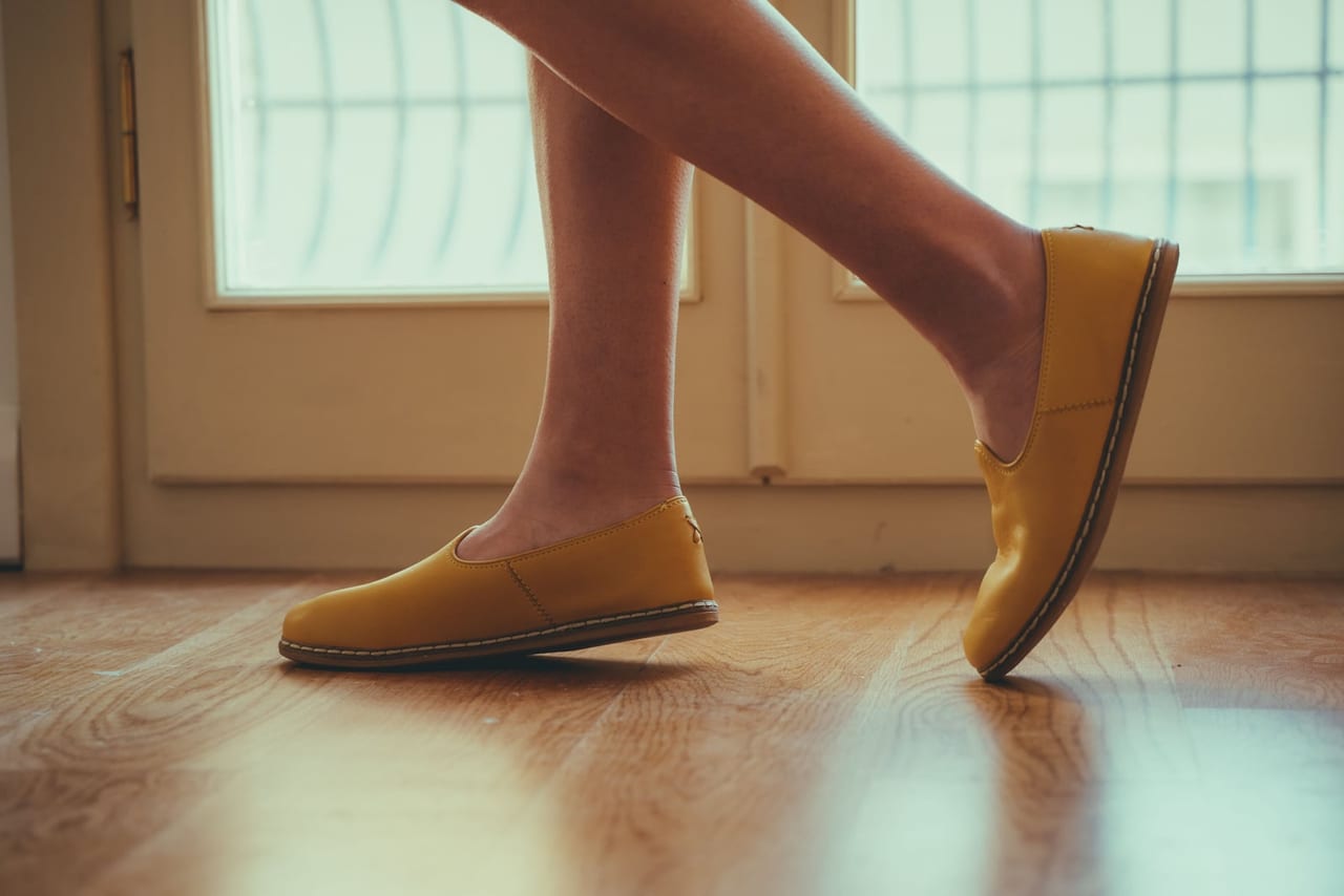 Bare legs in yellow slip-on shoes mid-stride on a wooden floor beside a window