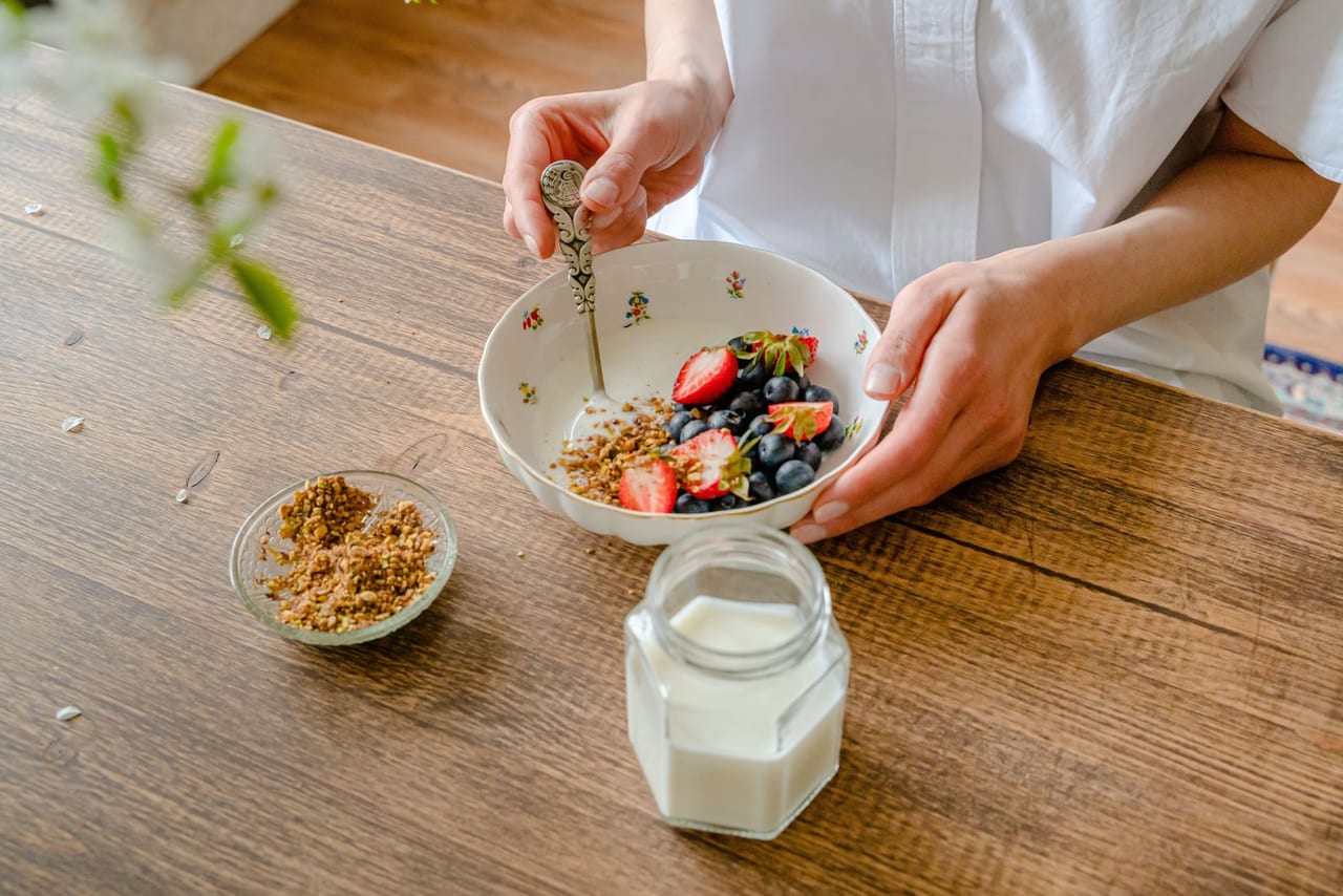 Hands around a bowl of yogurt with berries and granola beside a jar of milk